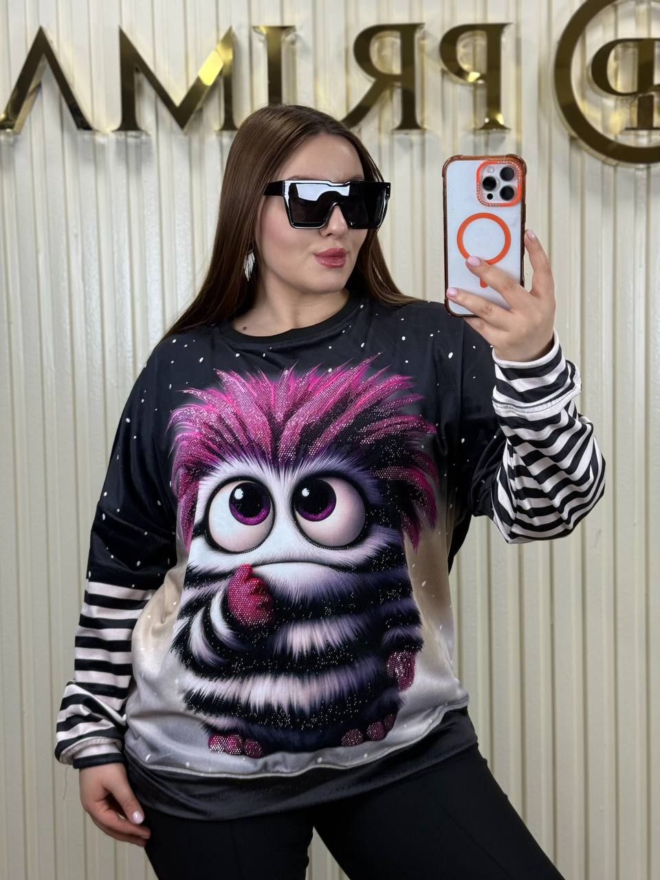 Person wearing a hoodie with a cartoon character design, taking a selfie in front of a branded backdrop.