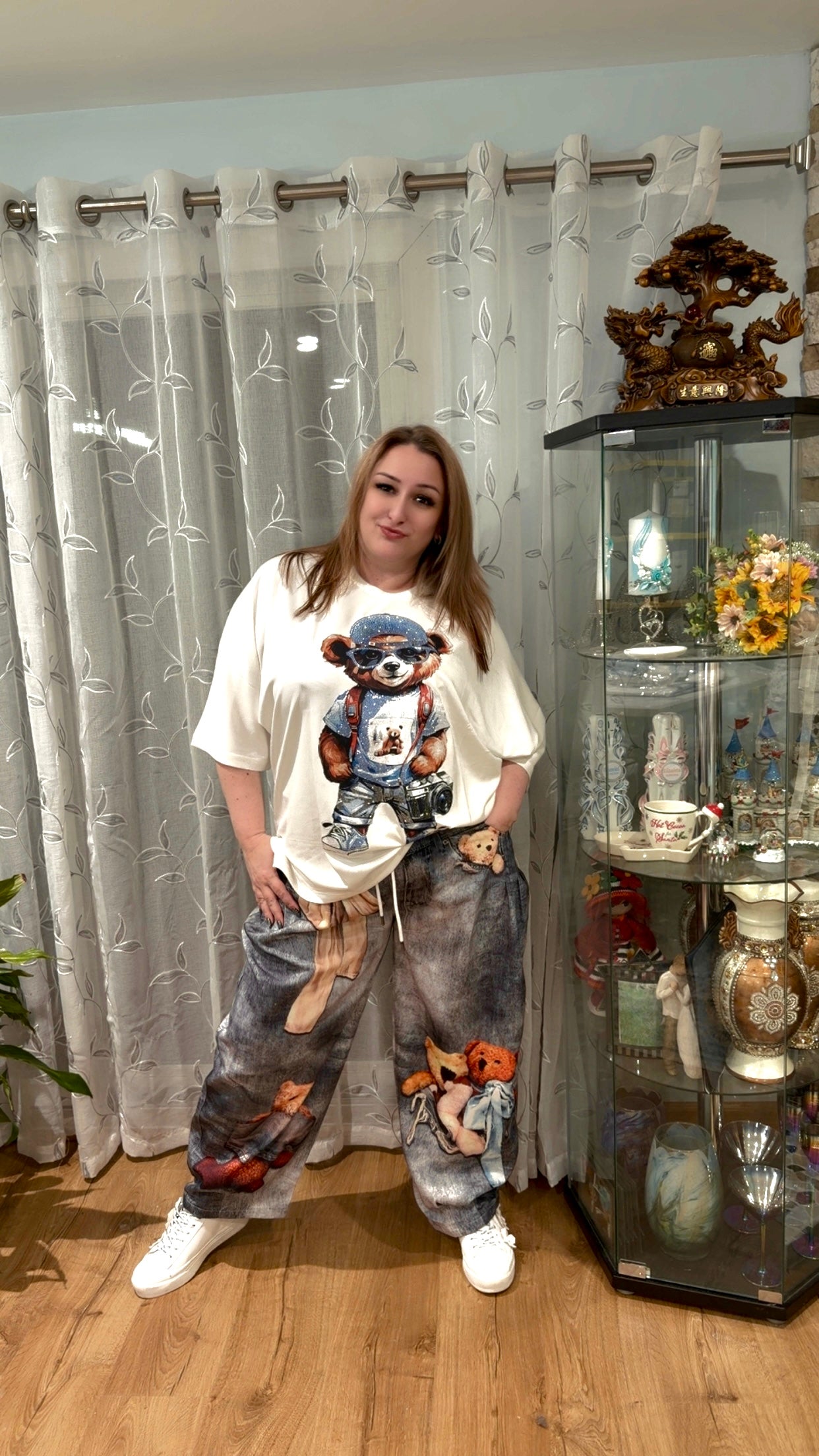 Person wearing a white t-shirt with a graphic design and gray pants standing in a room with curtains and a display cabinet.