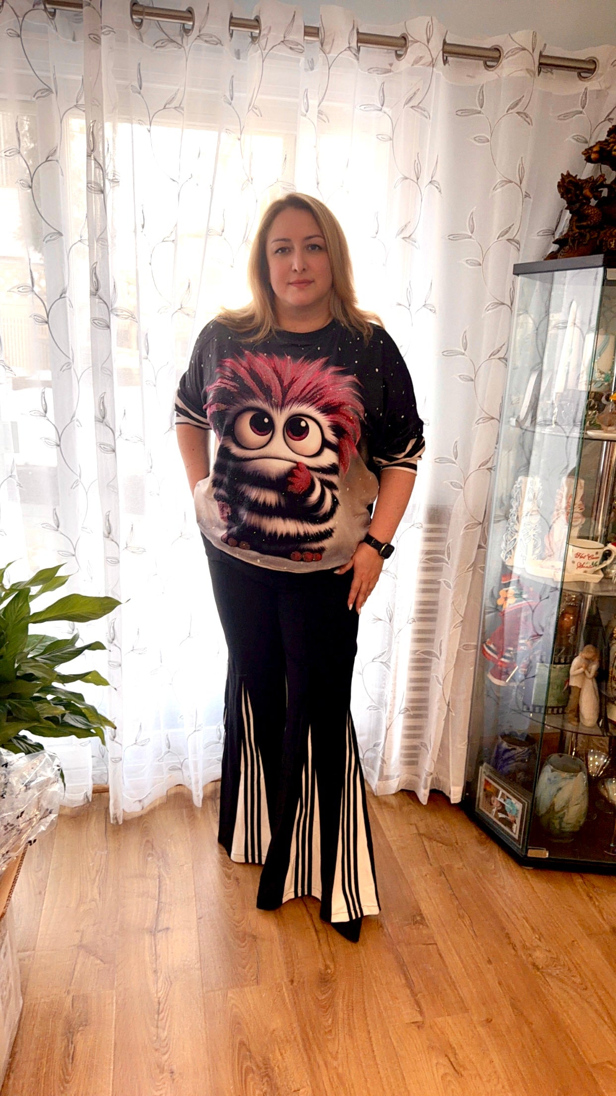 Person wearing a graphic t-shirt with a colorful design in a room with curtains and a plant.