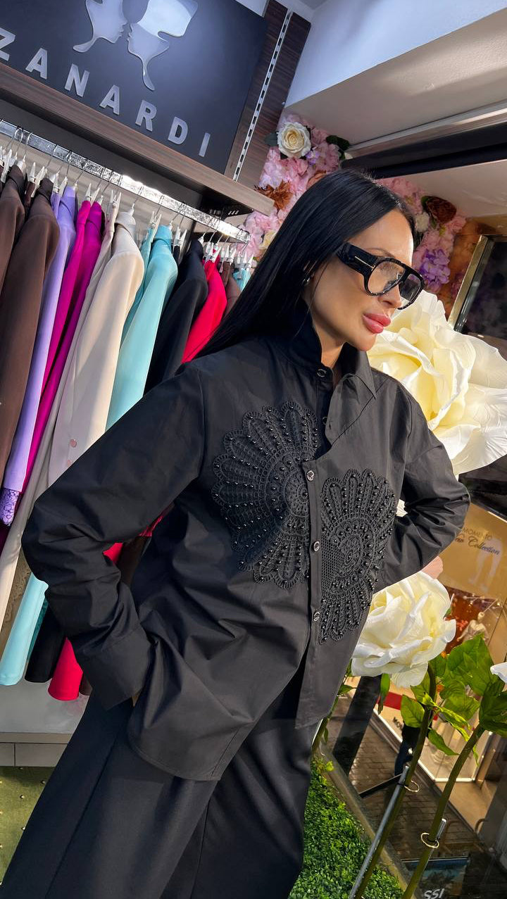 Woman in a black outfit with floral details standing in a store with clothing and decorative flowers.